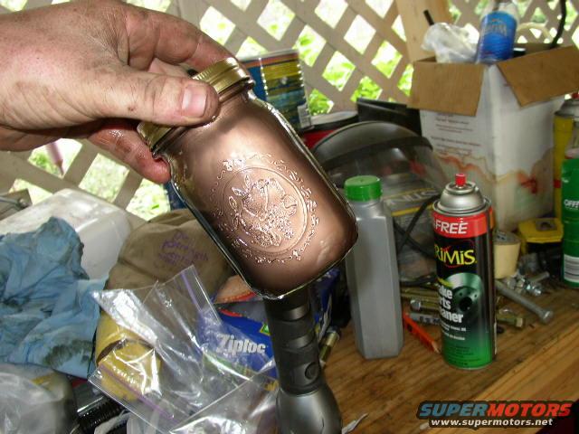Hand holding a copper-colored mason jar with embossed design. Background shows a workbench with tools and spray cans
