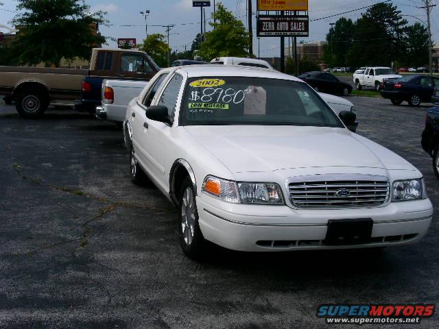 White Ford Crown Victoria with a "2002 $8780 LOW MILEAGE" sticker on the windshield, parked on asphalt