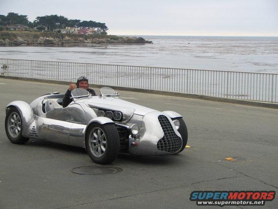 guy in a silver custom roadster giving a thumbs up, driving on a road by the ocean