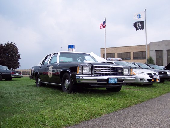 Front view of a black and white police car with a blue light bar on top. License plate reads '1979'. Other cars are parked behind it