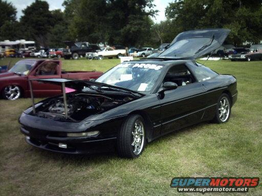Black 1995 Ford Probe with hood and rear hatch open, parked on grass at an outdoor event. Other vehicles visible in background