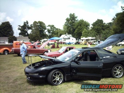 Black sports car with hood and trunk open at an outdoor car show. Other cars and people are visible in the background