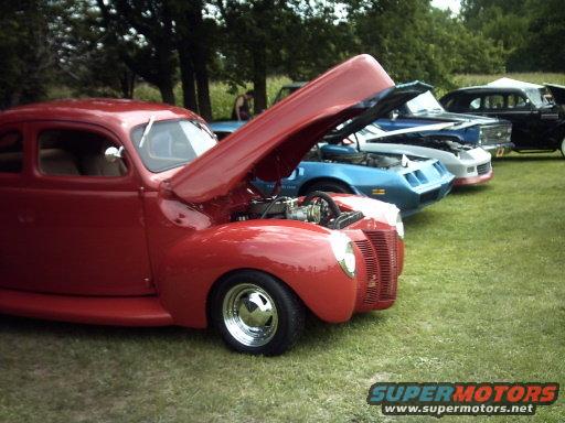 Red coupe with hood open, parked in a line of cars on grass