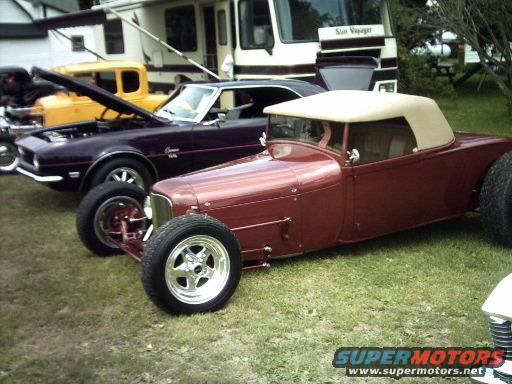 Maroon hot rod roadster with tan convertible top parked on grass next to a purple muscle car and a yellow truck