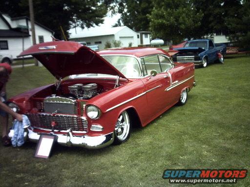 Red 1955 Chevrolet Bel Air coupe with hood open, chrome trim, and dual carburetors. A blue pickup truck is visible in the background