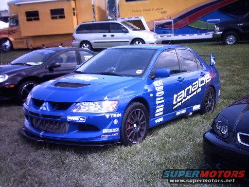 Blue Mitsubishi Lancer Evolution with 'tanabe' and other racing decals on the side, parked on grass. Black car to the left and front. Part of a yellow trailer and silver van in background