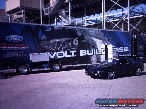 Black coupe parked in front of a large trailer with graphics. Trailer has Ford Racing branding and text that says 'VOLT. BUILD. RISE.'