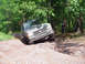 1979 Ford Bronco - An off-road vehicle tackles a challenging dirt track surrounded by trees, with a person observing in the background.