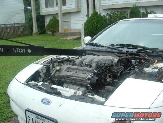 White car with hood open, engine exposed. Engine hoist above. Ford logo on front