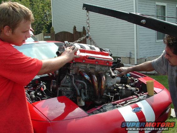 Two guys lowering a red-topped engine into the engine bay of a red car with white stripes. Ford badge visible on the car