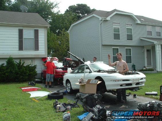 guy working on white car, engine out, red car behind it, parts everywhere