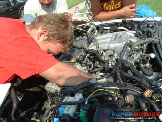 Engine bay with V6 engine being worked on by people, chain hoist attached