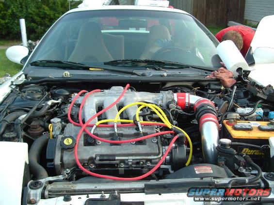 Engine bay of a white car with red and yellow spark plug wires, custom intake piping, and a Duracell battery