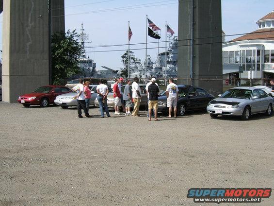 Cars parked near a waterfront with ships in the background. People are standing around