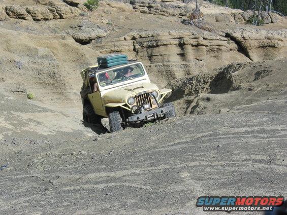 Yellow Jeep CJ-5 off-roading on a dirt slope with a green luggage carrier on the roof