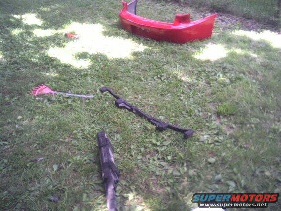 Red car bumper and yard tools on grass. Includes a rake and a black metal bar