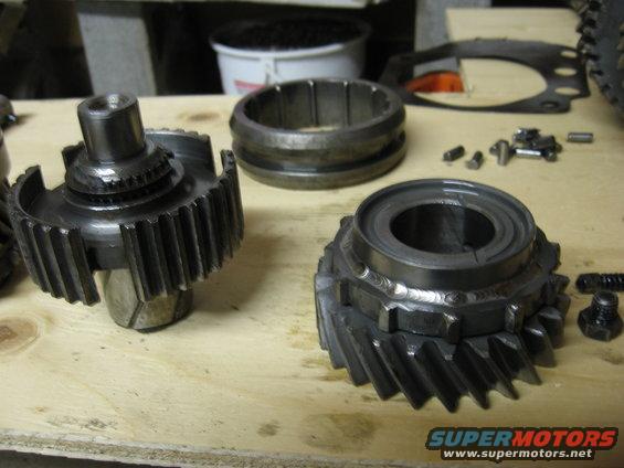 Transmission gears and synchronizer rings laid out on a wooden surface. Some small bolts and pins are also visible