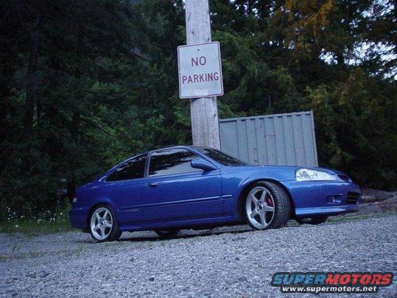 Blue coupe parked on gravel near a wooden post with a 'NO PARKING' sign