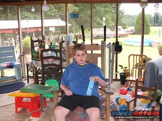 Young person in blue shirt and shorts sits in a rocking chair on a screened-in porch with various chairs and tables around