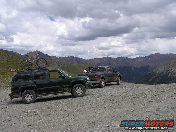 Dark green SUV with bicycle on roof rack, parked on a gravel road with a pickup truck behind it