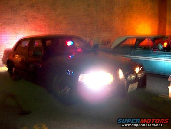 2001 Ford Crown Victoria police car with emergency lights illuminated, parked next to a light blue classic car