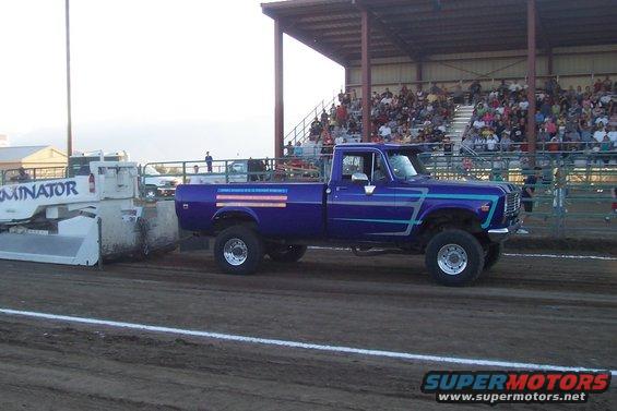 Purple pickup truck with blue racing stripes and an American flag decal on the bed, pulling a sled at a competition