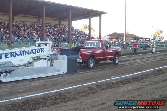 Red pickup truck pulling a sled down a dirt track with spectators in the stands