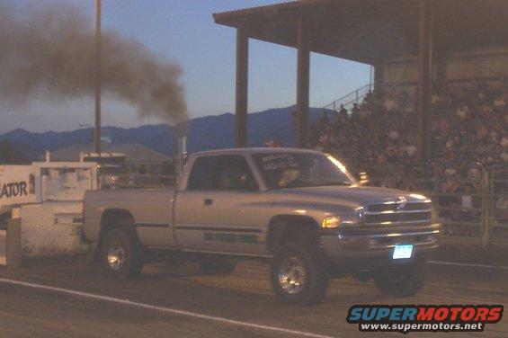Silver Dodge Ram pickup truck with a sled attached, emitting black smoke. Spectators in bleachers in background