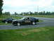 1978 Ford Mustang II - Two black Ford Mustang II Cobras are parked side-by-side on a gravel lot next to a road.