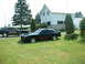 1978 Ford Mustang II - Two black 1978 Ford Mustang II Cobra II cars are parked on a lawn in front of a house. The car in the foreground has red side graphics.