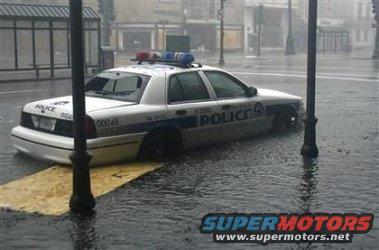 Police car submerged in floodwater, side view. The vehicle has "POLICE" text on its doors and emergency lights on the roof