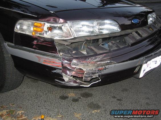 Dark sedan's front bumper is severely damaged and torn, with the left headlight assembly also impacted