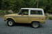 1972 Ford Bronco - Side profile of a tan and white 1972 Ford Bronco parked on a gravel surface with trees in the background.