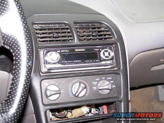Car dashboard with radio, air vents, and climate controls. Wires are visible in an open compartment below the controls
