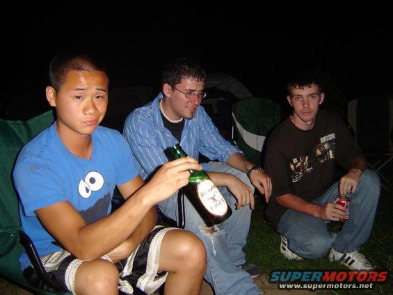 Three men sit in camping chairs at night, one holding a beer bottle