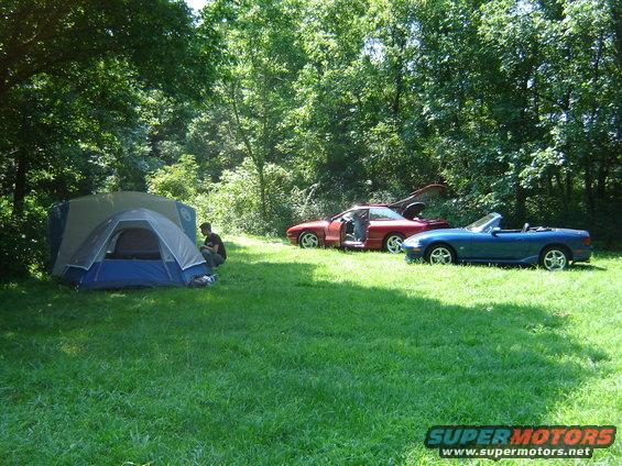 Red coupe with doors open and blue convertible parked in a grassy area with a tent
