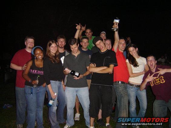 Group of young adults, some holding drinks, posing outdoors at night. One person holds up a trophy