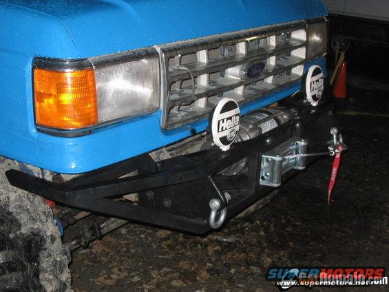 Blue Ford truck front with a chrome grille, orange turn signal, and a winch mounted on a black bumper. Two Hella lights are visible