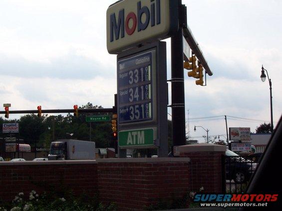 Gas station sign displaying gasoline prices and ATM, with traffic lights and a truck in the background