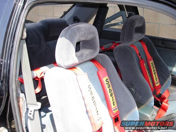 Car interior with gray racing seats and red Sabelt harnesses. Roll cage visible in background