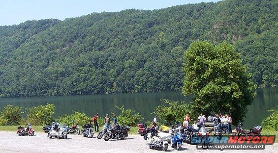 Group of motorcycles parked near a lake with a wooded hill in the background