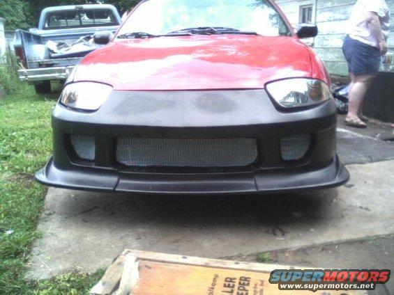 Front view of a red car with a black aftermarket bumper and mesh grille. A blue pickup truck is visible in the background