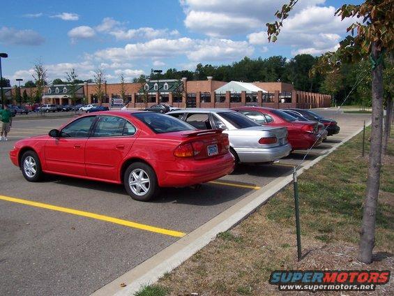 Red sedan parked in a lot next to other cars. The car has a spoiler and alloy wheels
