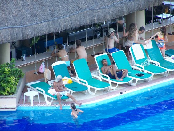 People relax on lounge chairs by a pool next to a thatched-roof bar