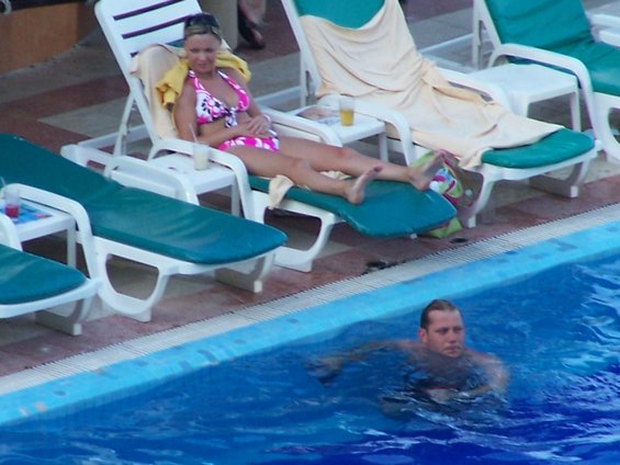 Woman in pink bikini on lounge chair by pool, man swimming