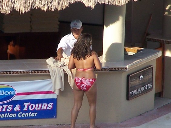 Woman in bikini talks to a resort worker behind a bar with 'SPORTS & TOURS' and 'TOWLAS/TOWELS' signs