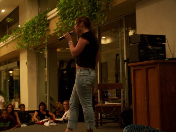 Woman in jeans and black top singing into a microphone indoors, with an audience seated in the background