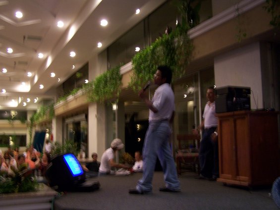 Man speaking into a microphone on a stage, with a monitor and audience in the background