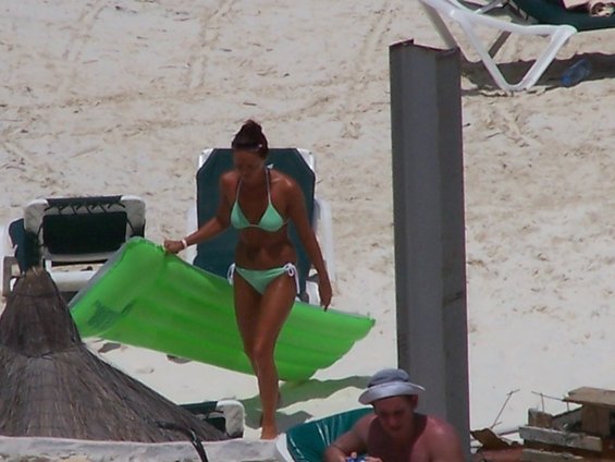 Woman in bikini carries a green inflatable mattress across a sandy beach with lounge chairs and a thatched umbrella visible