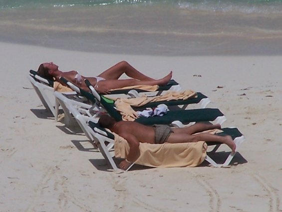 Two people relax on lounge chairs on a sandy beach with the ocean in the background